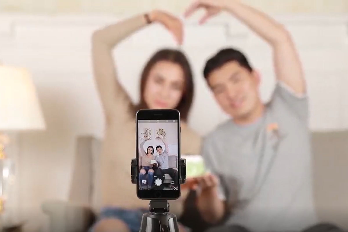 A selfie stick in the foreground, with a phone taking a photo of two people sitting on a couch with their arms making a heart shape