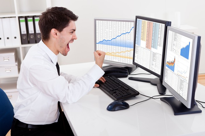 A stock investor pumping his fist as he looks at rising charts on his computer screens.