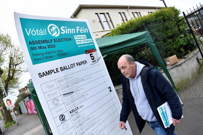 On May 5, 2022, in Belfast, Northern Ireland, a Sinn Fin activist stands outside a polling station in front of a sample ballot showing nationalists choosing whom to vote for. 